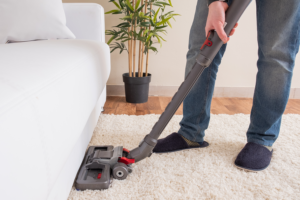 Man in blue jeans and navy slippers vacuuming white carpet in front of white couch and next to bamboo plant to improve indoor air quality.