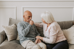 A senior man coughing into his hand while sitting on a grey couch next to his senior wife.