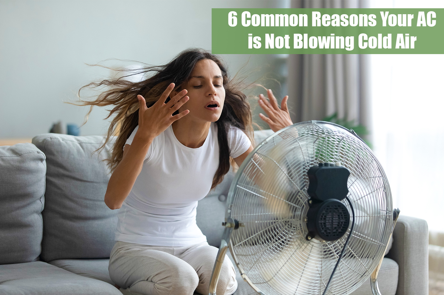 A woman in a white tshirt and long brown hair sitting on a gray couch in front of a fan because her AC is not blowing cold air.